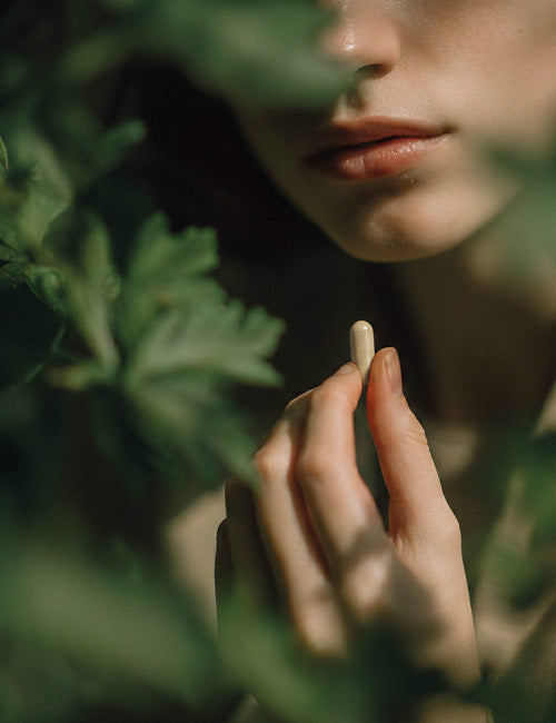 Kobieta trzymająca kapsułkę ashwagandhy w dłoni na tle zielonych roślin w naturalnym świetle