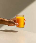 Dłoń trzymająca szklankę z pomarańczowym napojem kolagenowym na jasnym tle.