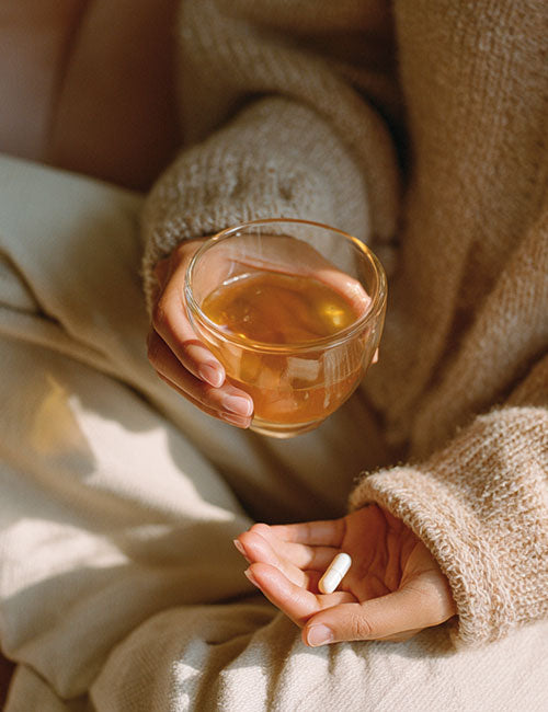 Osoba trzymająca kapsułkę suplementu Cynk Complex+ Biowen w dłoni oraz kubek z herbatą, przygotowująca się do suplementacji.