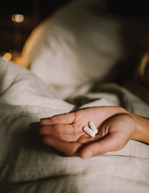 Dłoń trzymająca dwie białe kapsułki suplementu Glicyna Biowen na tle pościeli w ciepłym świetle.