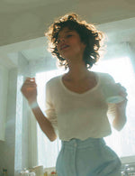 Kobieta w białej bluzce i niebieskich spodniach trzyma kubek przy oknie w kuchni.