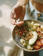 Zbliżenie na dłoń z kapsułkami suplementu Laktoferyna 96% Biowen i miskę pełną pestek, warzyw, awokado oraz pomidorów.