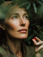 Zbliżenie twarzy kobiety otoczonej liśćmi, trzymającej czerwoną kapsułkę oleju z kryla.