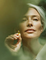 Kobieta trzymająca w dłoni żółtą kapsułkę suplementu Omega 3 Forte wśród rozmytej zieleni.