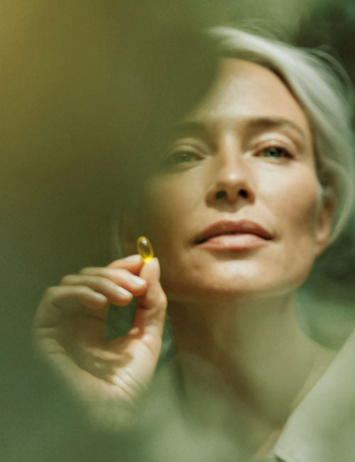 Kobieta trzymająca w dłoni żółtą kapsułkę suplementu Omega 3 Forte wśród rozmytej zieleni.