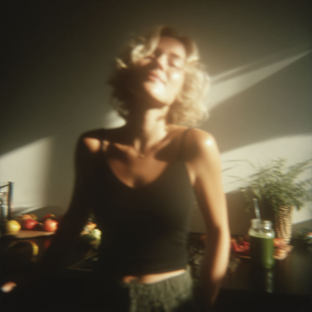 Kobieta w czarnym topie stoi w słońcu przy stole z napojem z Bio Chlorella Biowen 120 g i owocami.