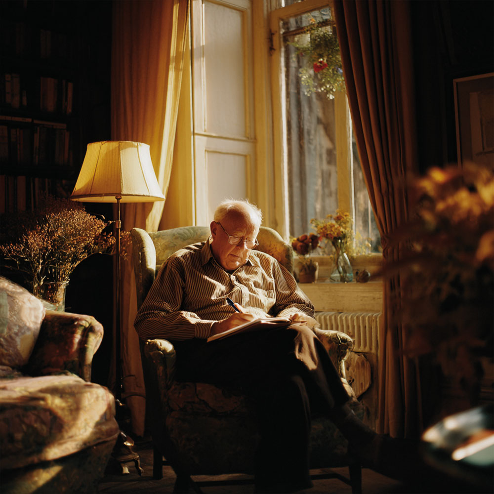 Starszy mężczyzna siedzi w fotelu przy oknie i pisze w notesie w ciepło oświetlonym pokoju.