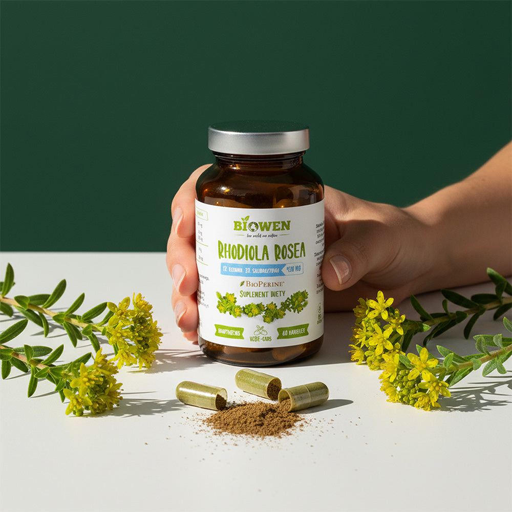Ręka trzyma słoiczek suplementu Rhodiola Rosea (różeniec górski) Biowen 60 kapsułek obok kwiatów i rozsypanego proszku z kapsułek.