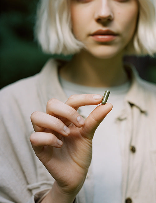 Kobieta o jasnych włosach trzyma w palcach zieloną kapsułkę suplementu Rhodiola Rosea (różeniec górski) Biowen.