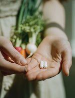 Zbliżenie na dłoń z dwiema kapsułkami Biowen Witamina B12 Complex+ i bukietem świeżych warzyw w tle.