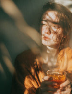 Kobieta w brązowym swetrze trzymająca szklankę z napojem w ciepłym świetle z cieniami liści na twarzy.