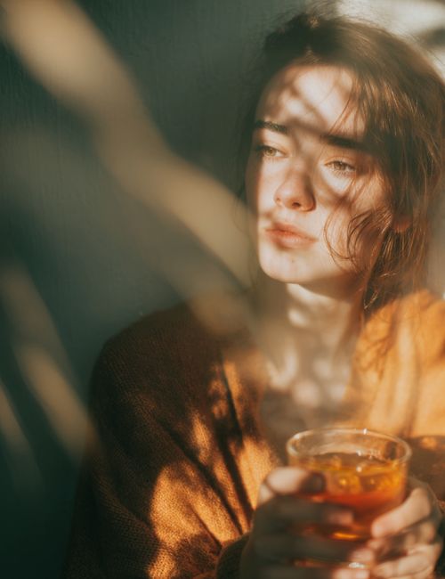 Kobieta w brązowym swetrze trzymająca szklankę z napojem w ciepłym świetle z cieniami liści na twarzy.