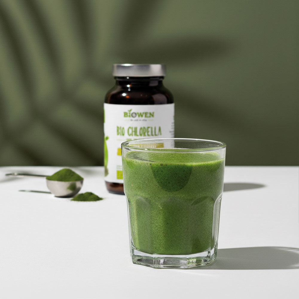 Szklanka zielonego napoju z Bio Chlorella Biowen obok słoiczka suplementu i łyżki proszku.