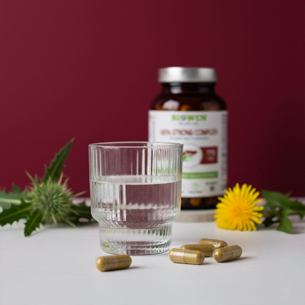 Szklanka wody i kapsułki Hepa Strong Complex+ Biowen na stole z butelką suplementu i kwiatami w tle.