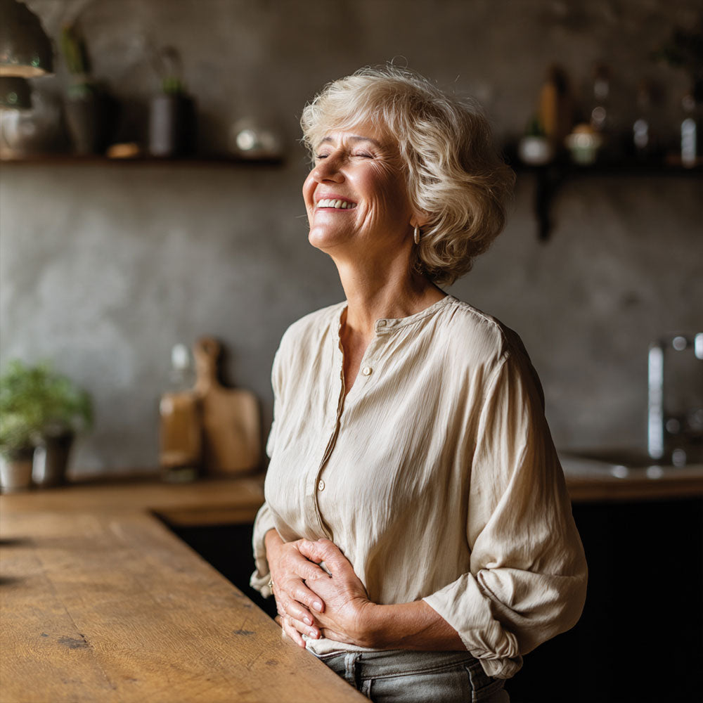 Uśmiechnięta kobieta w beżowej koszuli opiera się o blat kuchenny, trzymając się za brzuch.