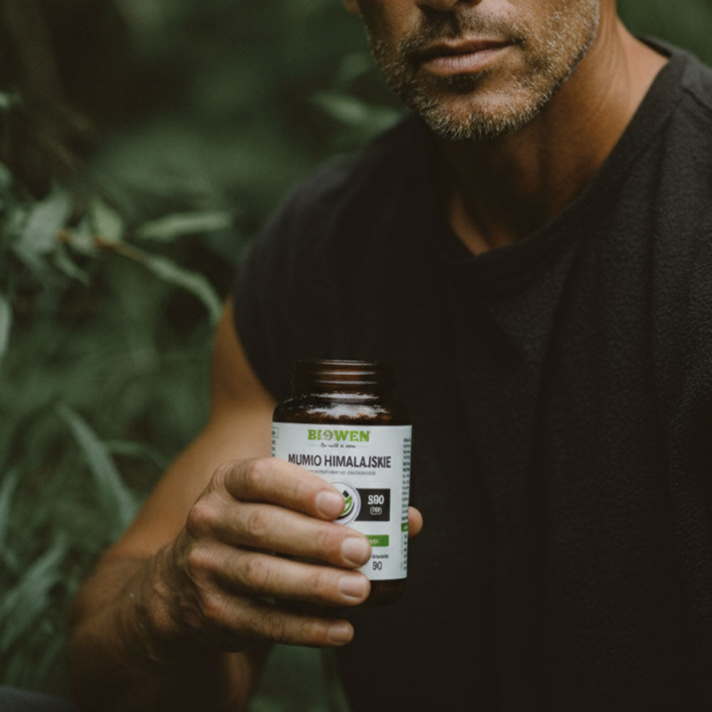 Mężczyzna w czarnej koszulce trzymający słoik suplementu Mumio himalajskie Biowen 90 kapsułek wśród zieleni.