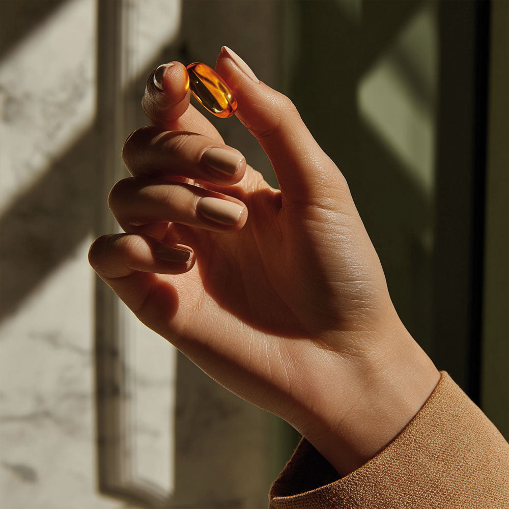 Dłoń trzymająca bursztynową kapsułkę suplementu Omega 3 w świetle dziennym.