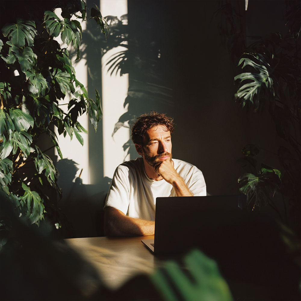 Mężczyzna siedzący przy biurku z laptopem wśród roślin, oświetlony naturalnym światłem.