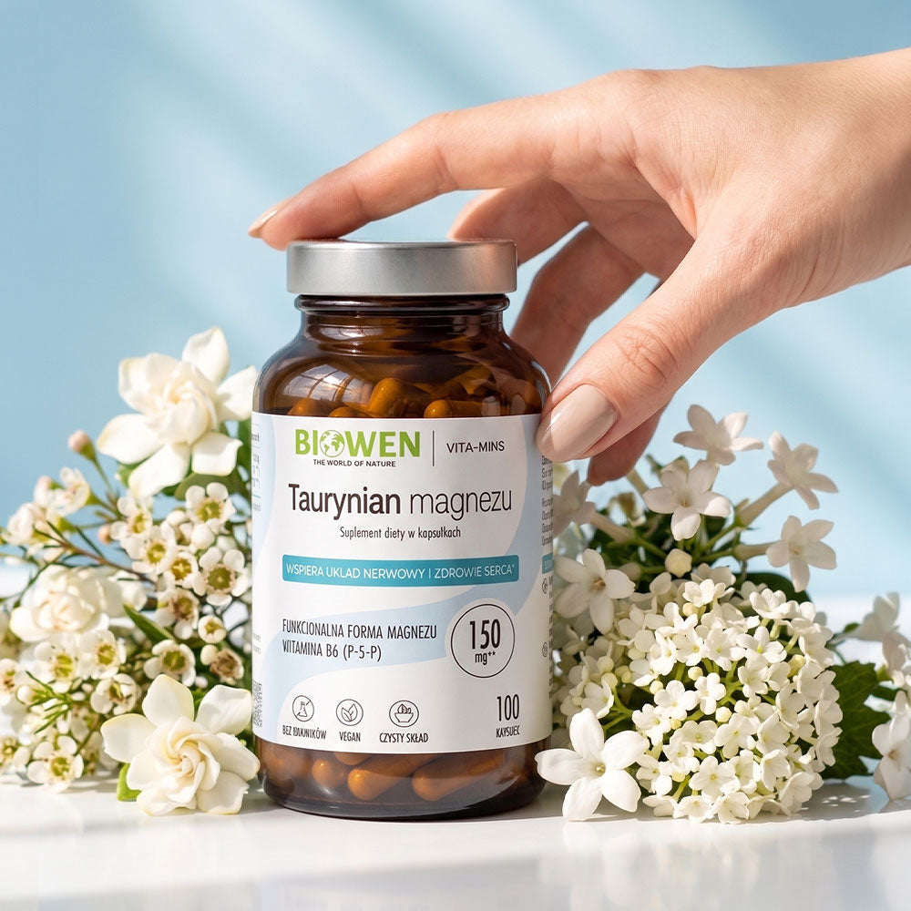 Dłoń sięgająca po Biowen Taurynian Magnezu 150 mg z witaminą B6, 100 kapsułek, wśród białych kwiatów.