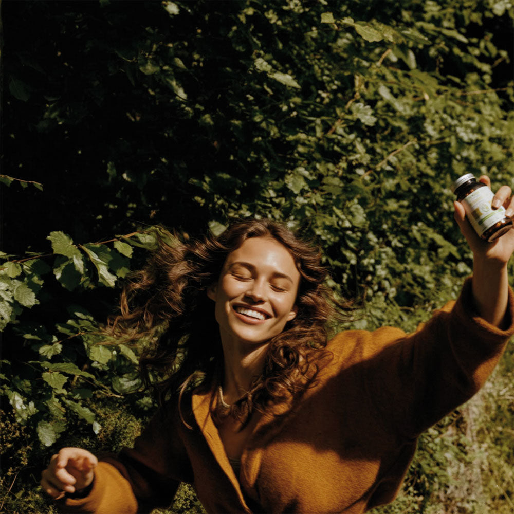 Kobieta w brązowym płaszczu trzymająca Witamina D3 Biowen 120 kapsułek na tle zielonych liści.