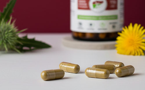 Kapsułki suplementu Hepa Strong Complex+ Biowen na stole z ostropestem i mniszkiem lekarskim w tle.