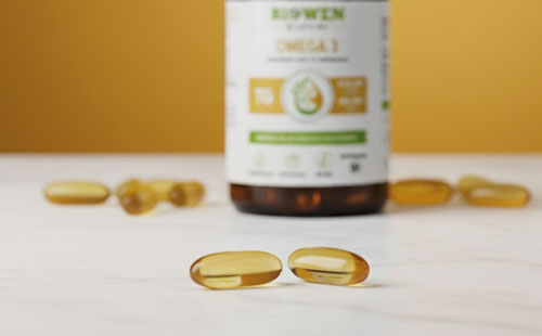 Kapsułki Omega 3 Biowen ułożone na jasnej powierzchni przed brązową butelką suplementu.