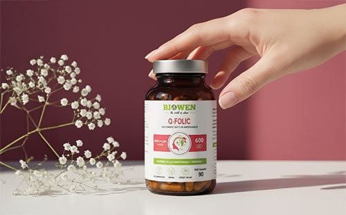 Ręka sięgająca po suplement Q-folic Kwas Foliowy Biowen 90 kapsułek na tle różowej ściany z gałązką gipsówki.