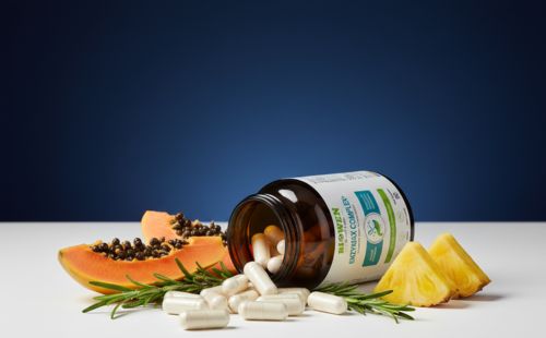 Przewrócony słoiczek Biowen EnzyMax Complex+ z kapsułkami obok papai i ananasa