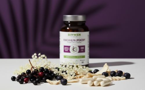 Butelka Biowen Kwas R-Alfa-Liponowy otoczona kapsułkami, owocami aronii, czarnego bzu i kwiatami bzu na jasnym blacie.
