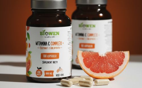 Dwa słoiczki Biowen Witamina C Complex+ 100 kapsułek z kapsułkami i kawałkiem grejpfruta na pomarańczowym tle.