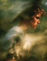 Mężczyzna w jasnej koszulce stojący wśród rozmytych, zielonych roślin w miękkim świetle.