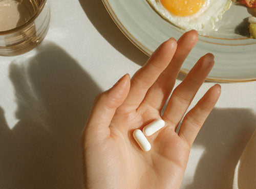 Dłoń z dwiema kapsułkami Jabłczanu magnezu Biowen obok talerza z jajkiem sadzonym.