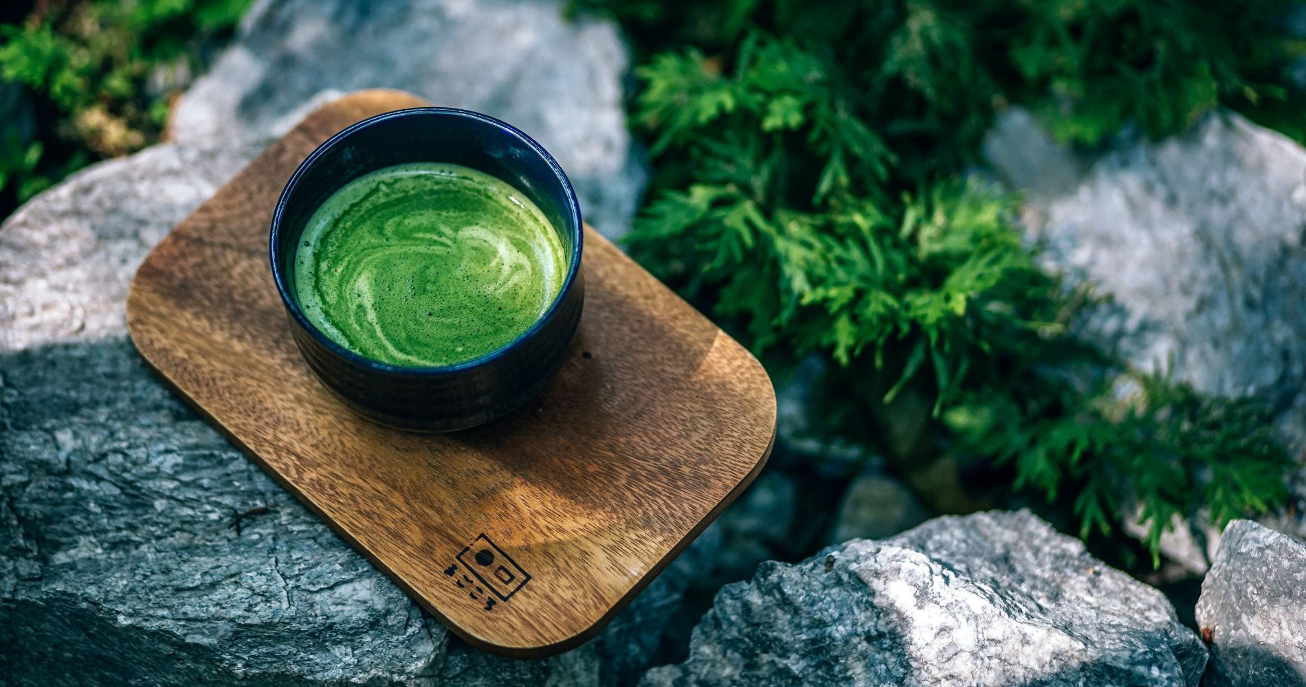 Filiżanka zielonej herbaty matcha na drewnianej podstawce, w otoczeniu kamieni i roślin.